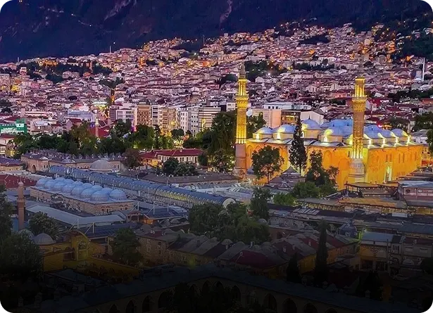 Aerial night view of Bursa Grand Mosque illuminated with golden lights against the city and Uludağ mountain backdrop