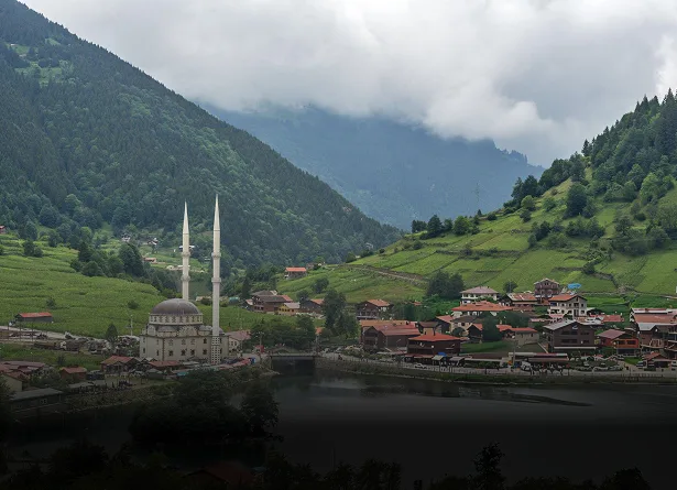 Mosque with twin minarets beside a river in a green mountain village in Trabzon, surrounded by mist-covered forested hills and red-roofed houses