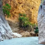 A narrow river flows between steep, rocky canyon walls with sparse green trees growing from the cliffs—a perfect setting for a Kayakoy Chill Out Day. The water is light blue and the rocks are beige and orange, creating a tranquil escape amidst nature’s beauty.